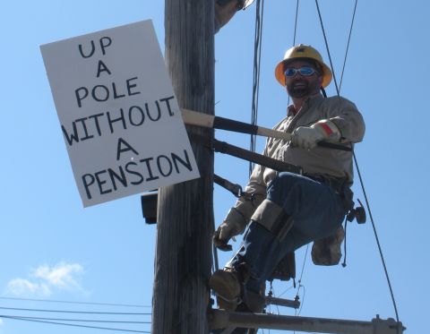 Utility worker climbing a pole beside a protest sign by IBEW 1837