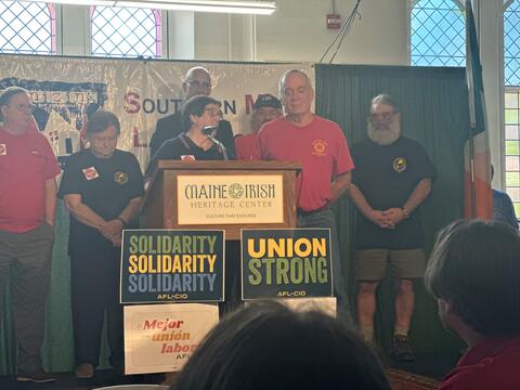 Speaker at podium with union solidarity signs by IBEW 1837