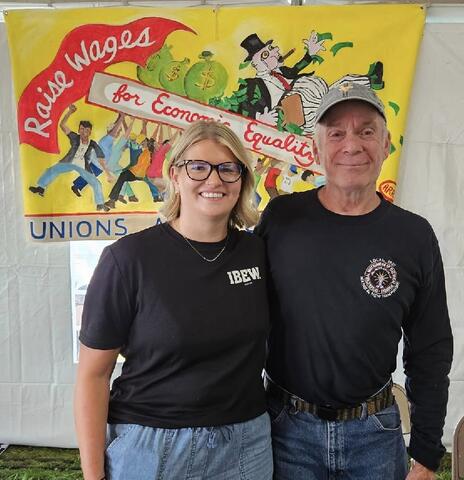Two IBEW members posing in front of a union fair banner by IBEW 1837